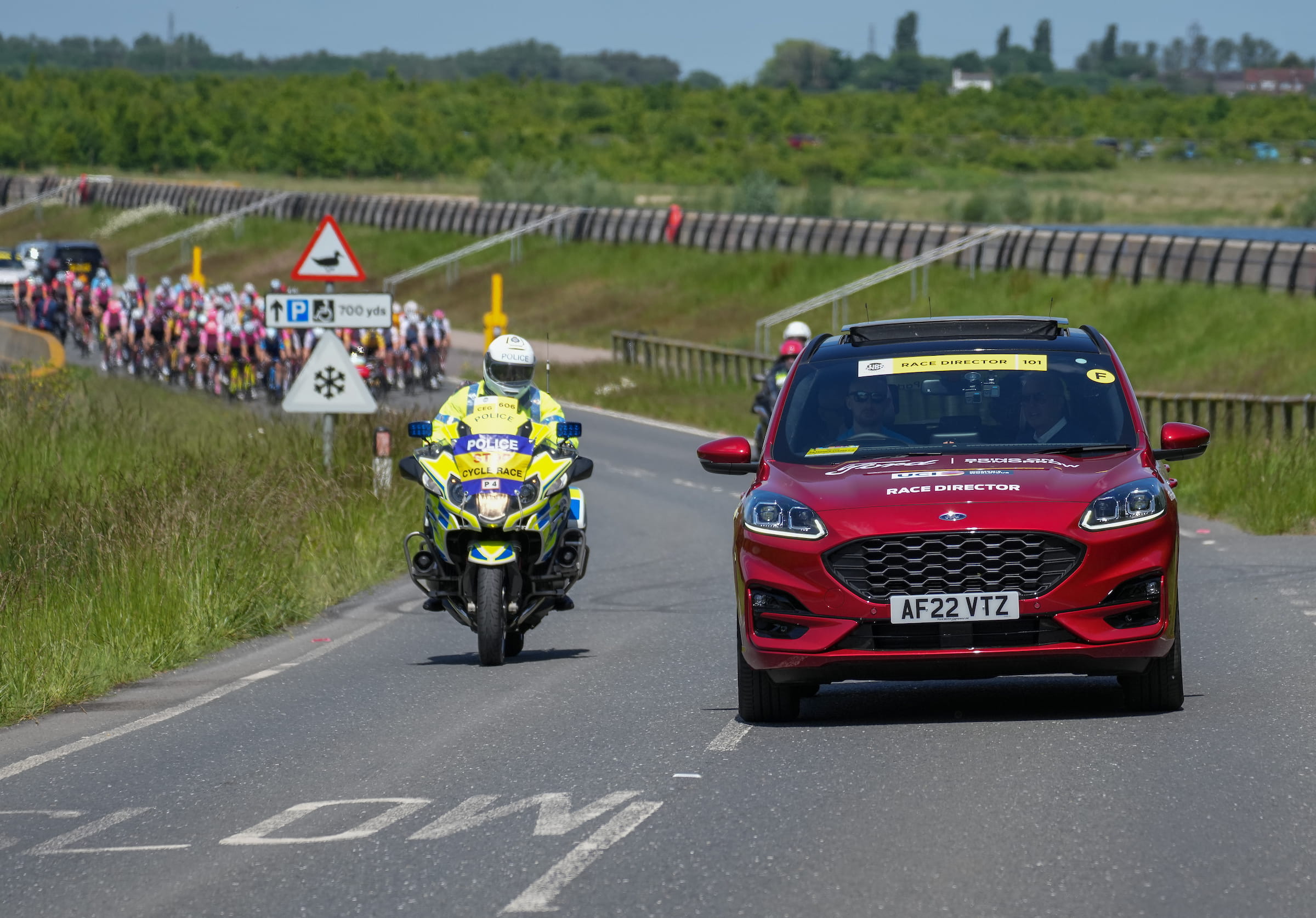 A police motorcycle and car drive ahead of a peloton of cyclists